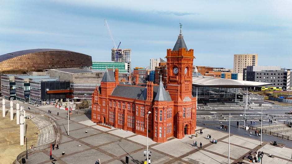 A waterfront scene in Cardiff Bay featuring a large white Ferris wheel situated behind a historic red-brick building with a prominent clock tower. In the foreground, the water reflects the structures