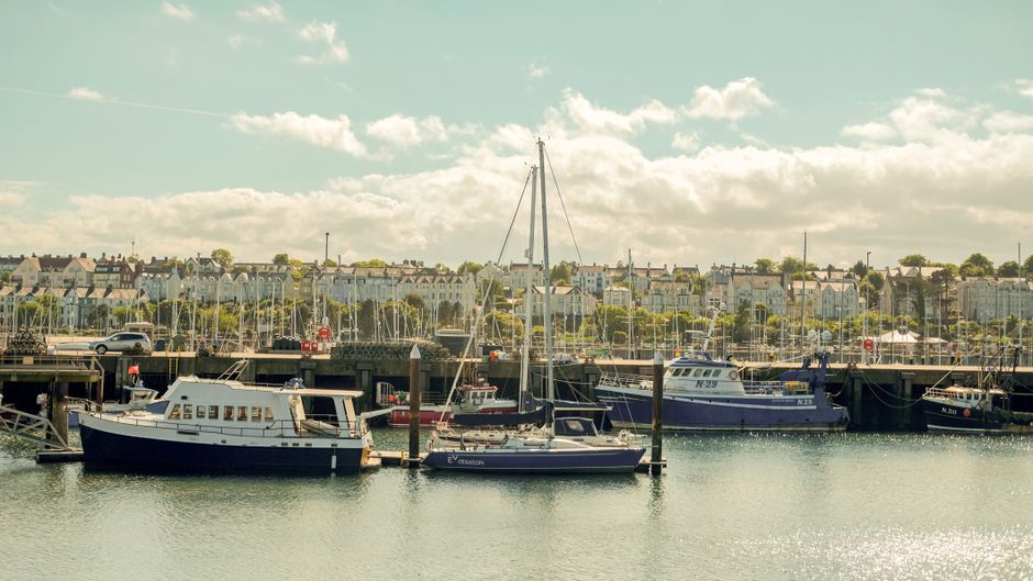 A view of a marina with several moored sailboats and motorboats, located alongside a paved quay. In the foreground, a rusted metal bollard connected to a large worn rubber tire with a rusty chain, whi